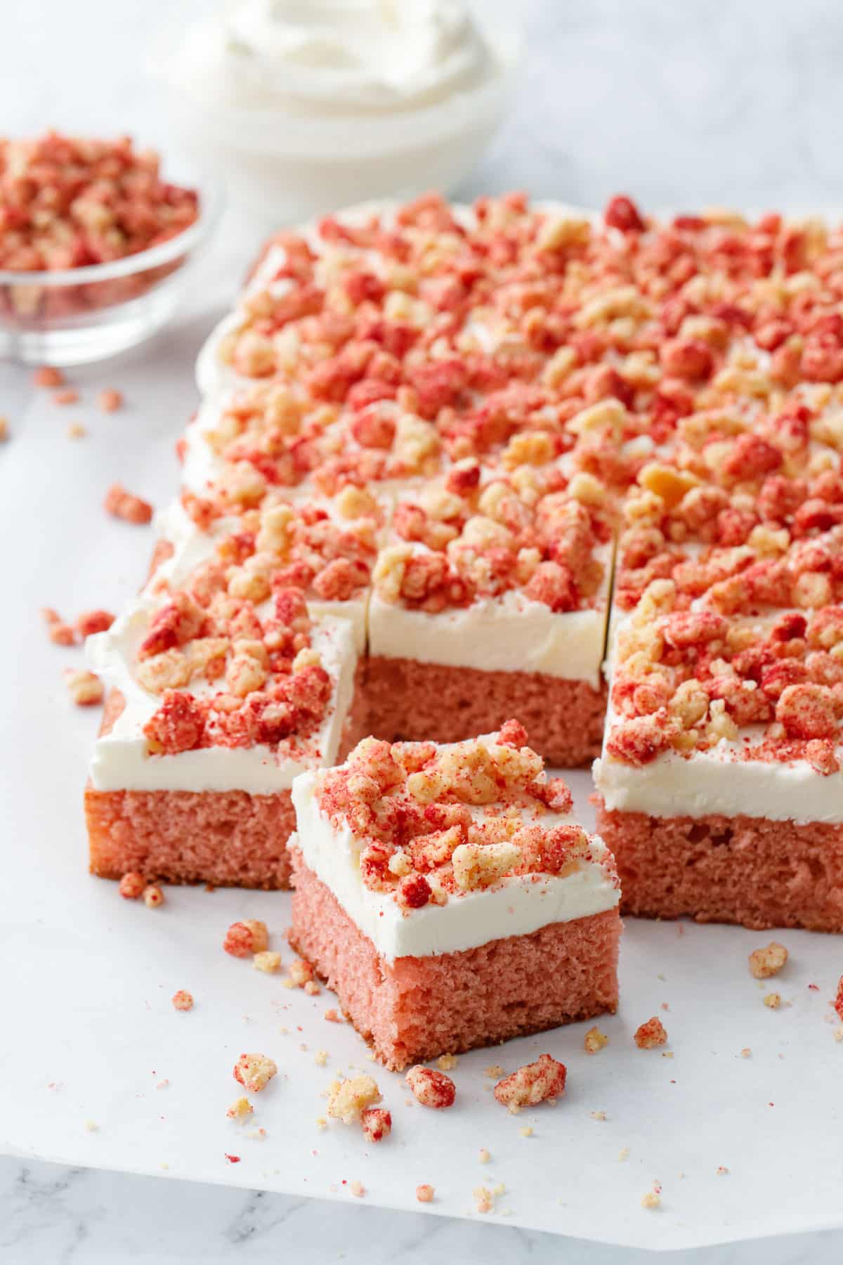 Strawberry Crunch Sheet Cake with Buttermilk Frosting cut into serving squares, on a piece of parchment with messy crumbs, and a bowl of more crumbs and frosting in the background.