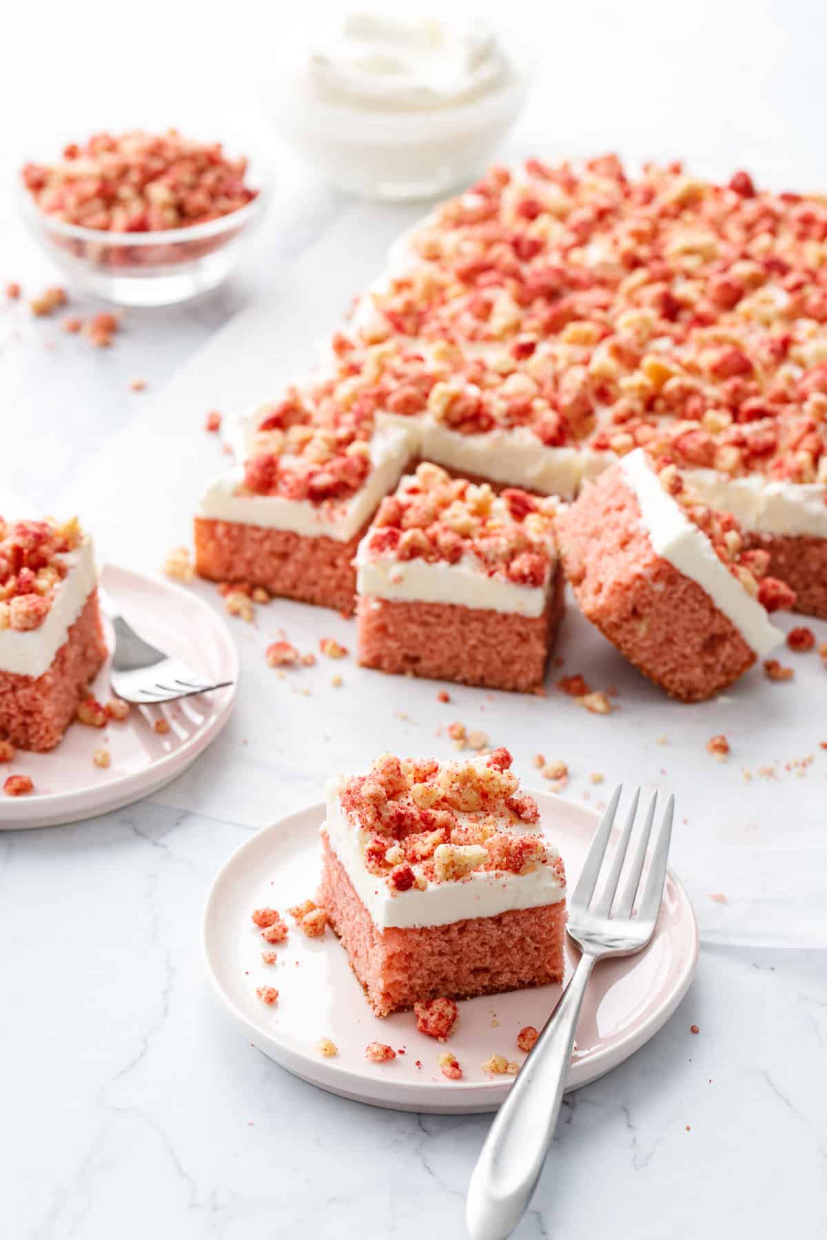 Strawberry Crunch Sheet Cake with Buttermilk Frosting, two cut squares on small dessert plates with forks, and a bowl of more crumbs and frosting in the background.