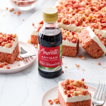 Strawberry Crunch Sheet Cake with Buttermilk Frosting, two squares on dessert plates with forks, focus on a bottle of Amoretti Wild Strawberry Artisan flavoring.