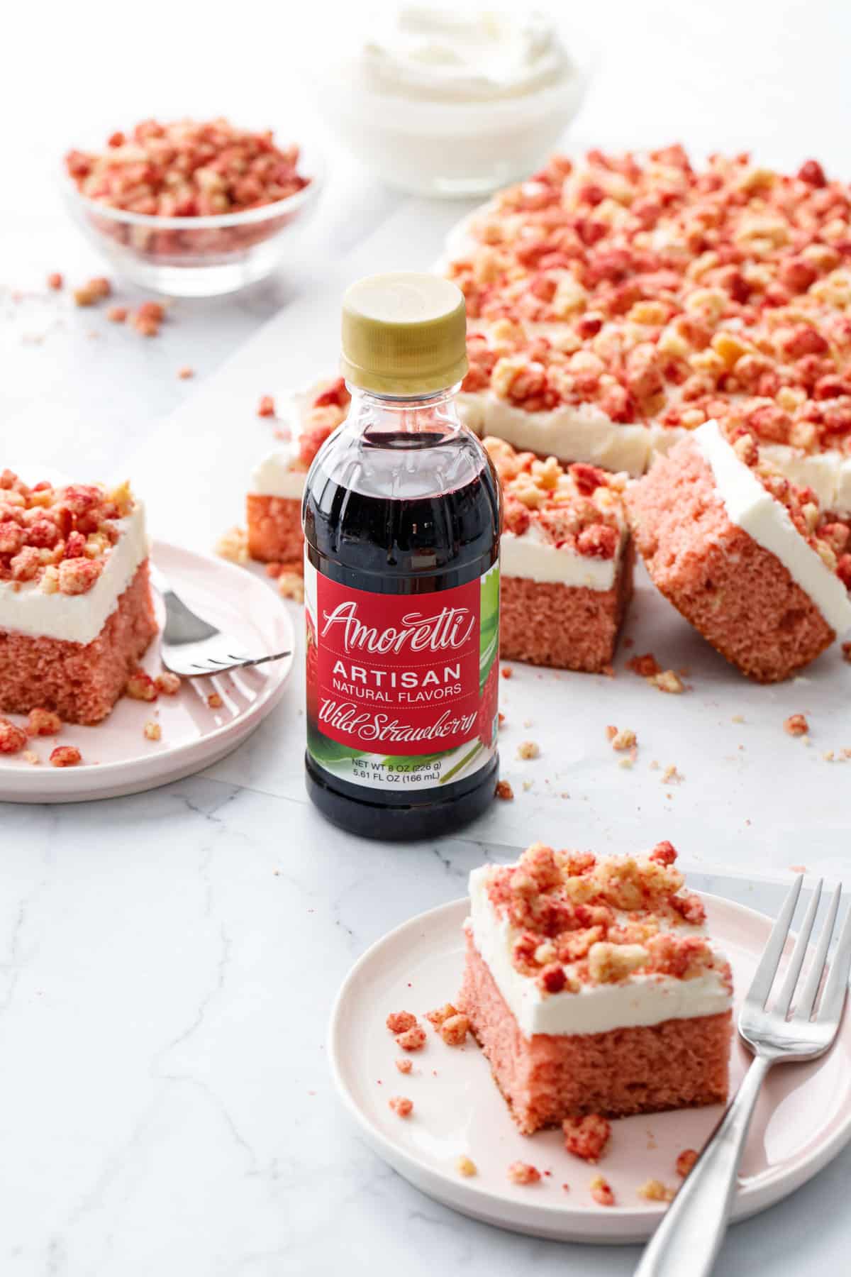 Strawberry Crunch Sheet Cake with Buttermilk Frosting, two squares on dessert plates with forks, focus on a bottle of Amoretti Wild Strawberry Artisan flavoring.