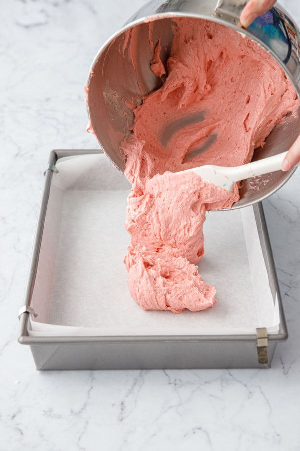 Pouring pink strawberry cake batter into a parchment-lined 13x9 metal baking pan.