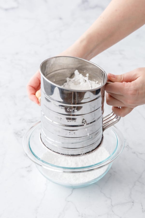 Metal sifter sifting powdered sugar into a glass mixing bowl.