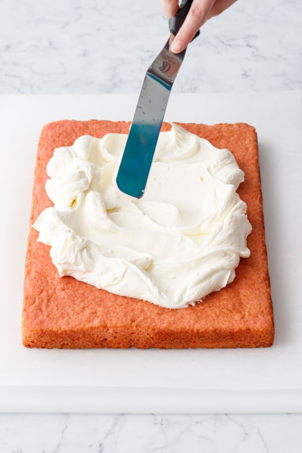Spreading fluffy white buttermilk buttercream onto a pink colored strawberry sheet cake sitting on a piece of parchment paper.