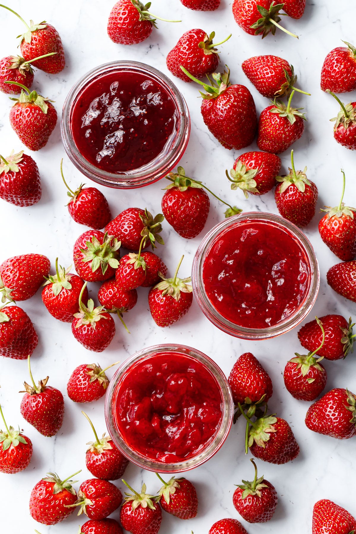 Overhead, marble background with lots of scattered fresh strawberries and three open jars of different kinds of strawberry jam.