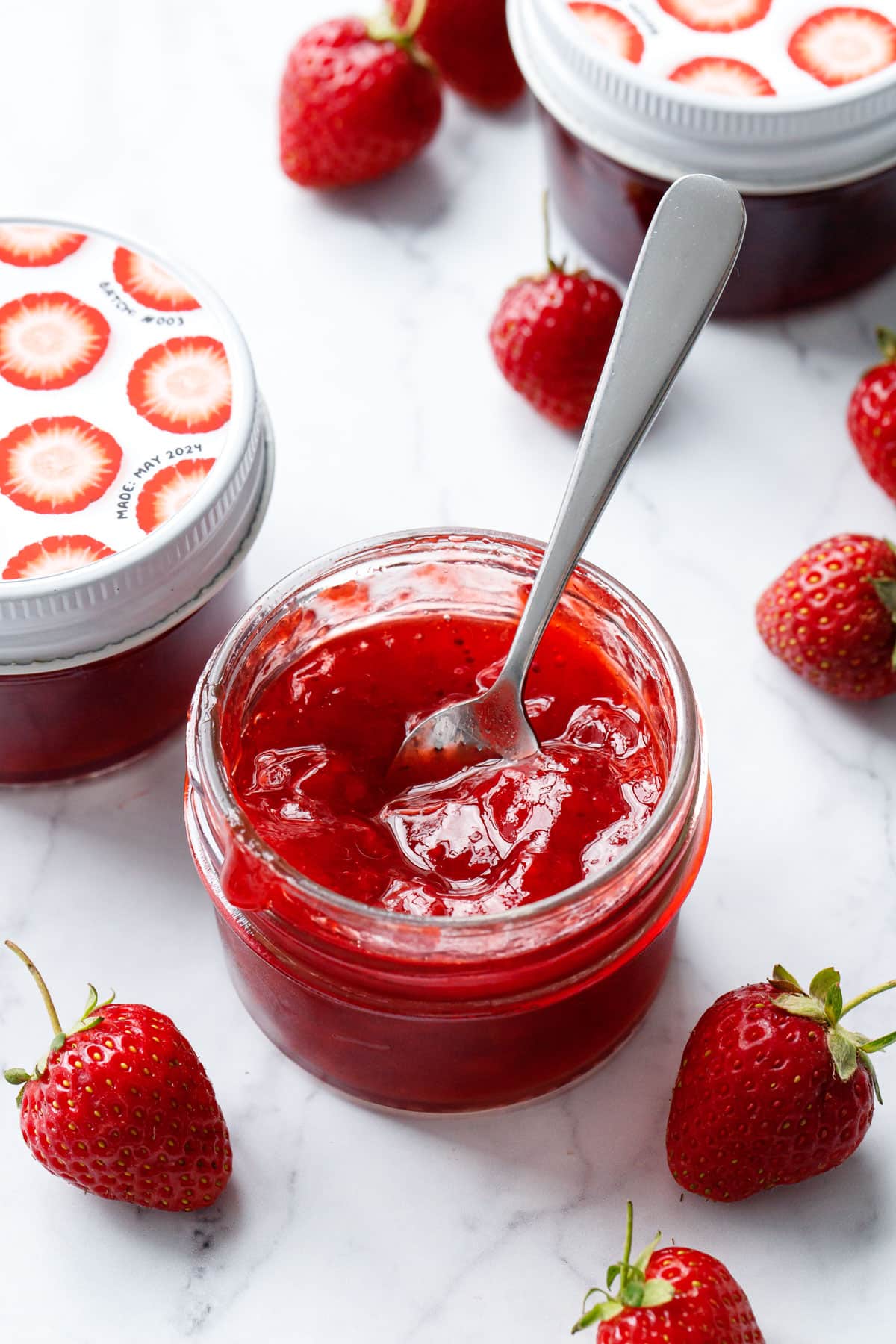 Classic Strawberry Jam in an open glass jar with a spoon to show texture, with more jars of jam and fresh strawberries in the background.
