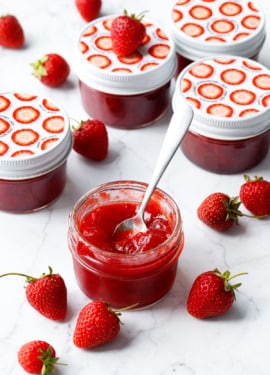 Classic Strawberry Jam in a glass jar with fresh strawberries on a marble background, one jar open in the foreground with a spoon in it to show the texture.