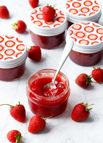 Classic Strawberry Jam in a glass jar with fresh strawberries on a marble background, one jar open in the foreground with a spoon in it to show the texture.
