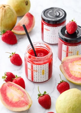 Jars of Guava Strawberry Jam on a marble background with fresh strawberries and whole and cut pink guavas scattered around.