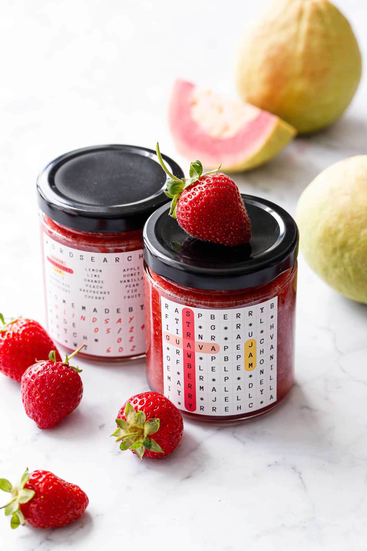 Two jars of Guava Strawberry Jam with black lids on a marble background with fresh strawberries and pink guavas in the background.