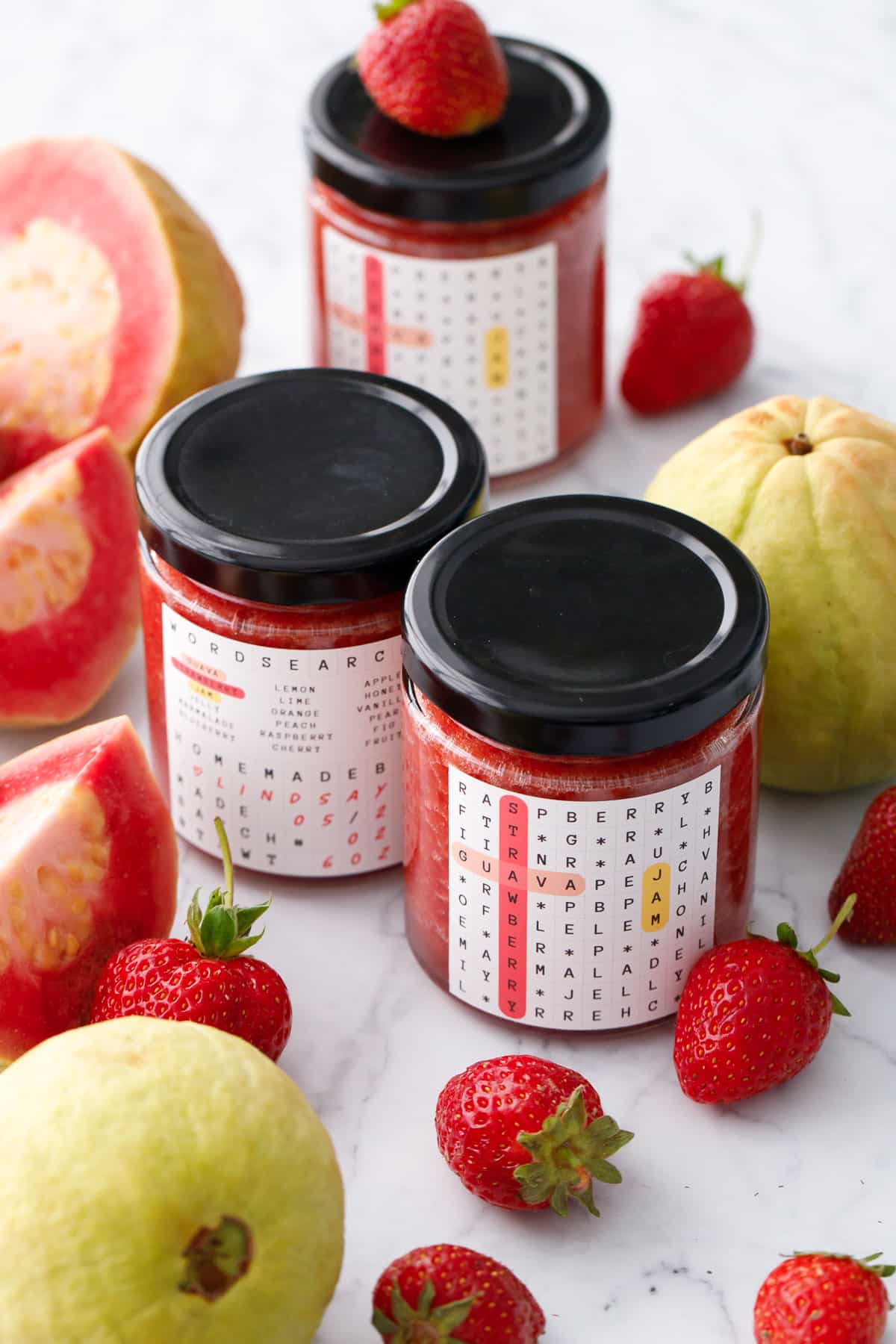 Three jars of Guava Strawberry Jam with black lids on a marble background with fresh strawberries and pink guavas in the background.