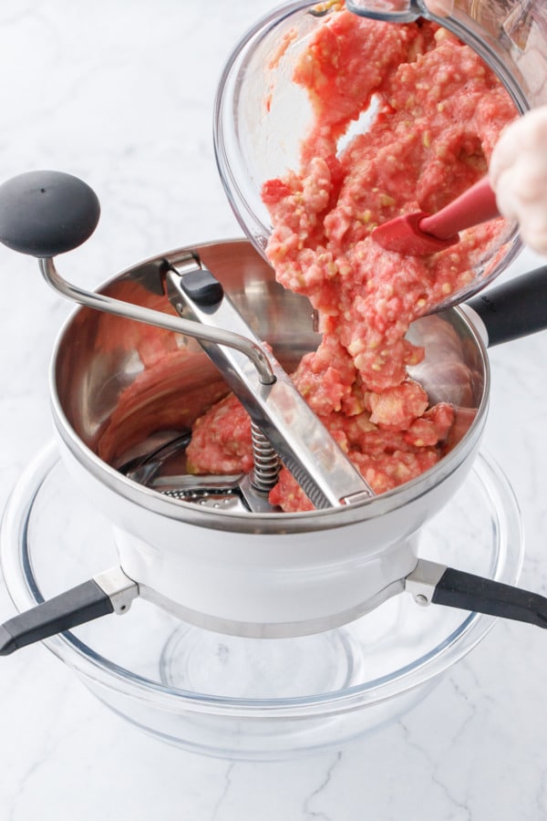 Pouring blended guava puree into a food mill set over a glass mixing bowl.