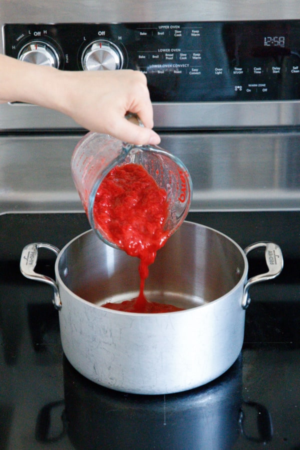 Pouring mashed strawberry into a brushed stainless saucepan on a glass stovetop.