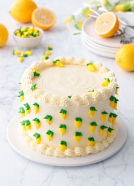 Triple Lemon Layer Cake decorated with tiny royal icing lemons, on a white cake plate with marble surface and lemons in the background.