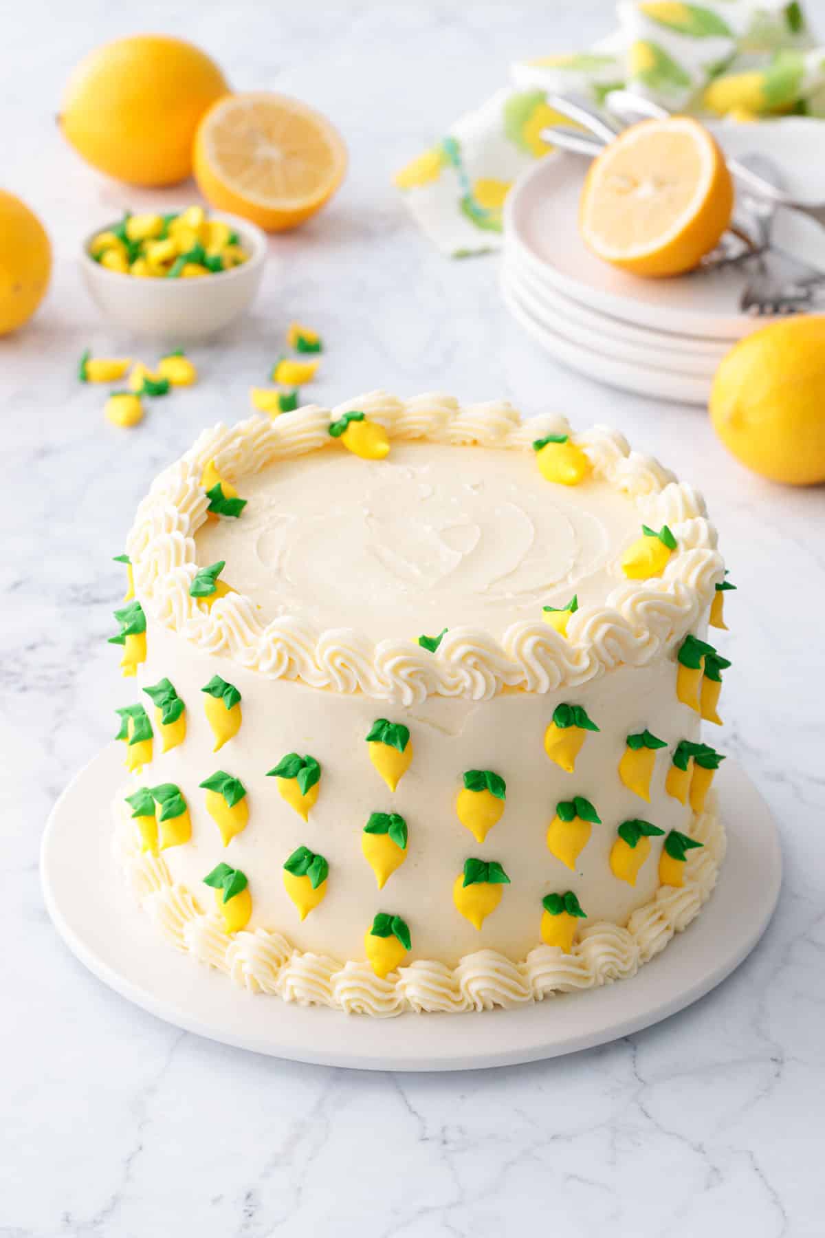 Triple Lemon Layer Cake decorated with tiny royal icing lemons, on a white cake plate with marble surface and lemons in the background.