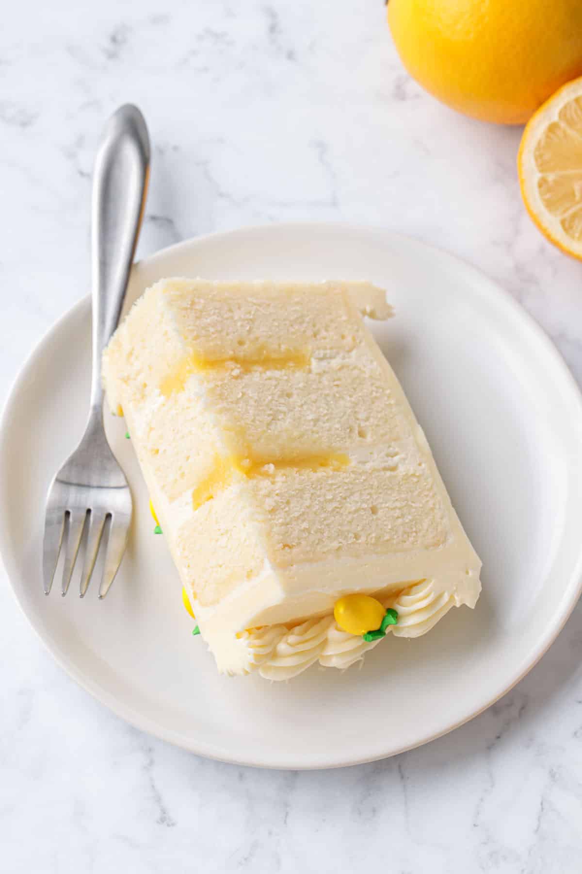 Slice of Triple Lemon Layer Cake on a white plate with fork and lemons off to the side.