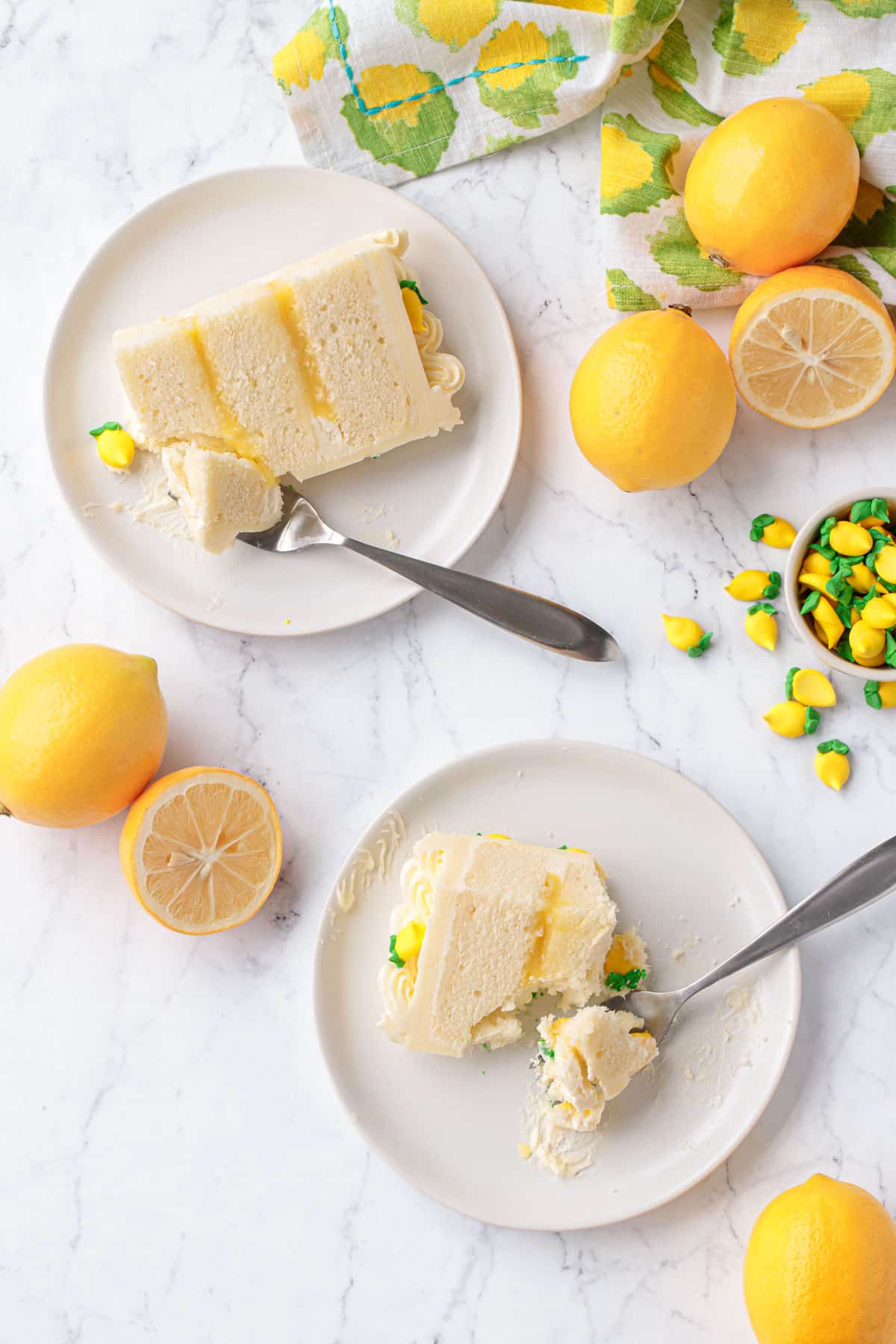 Overhead, two slices of Triple Lemon Layer Cake on white plates with forks and bites taken out, with some more lemons and little bowl of royal icing lemon decorations on the side.
