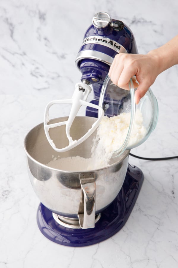 Pouring lemon zest-infused sugar into a mixing bowl with flour and other dry ingredients.