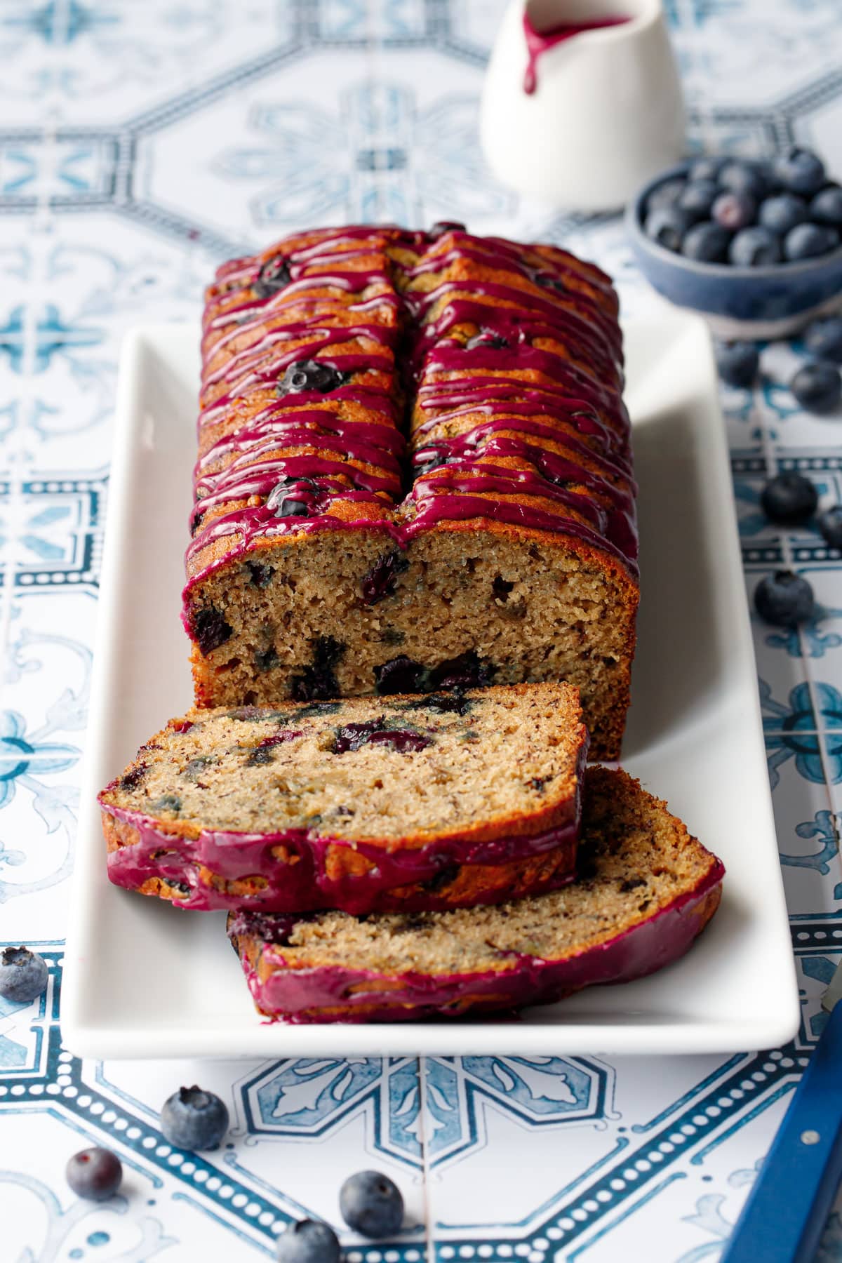 Loaf of Blueberry Banana Bread on a white rectangular plate with two slices lying down on a blue and white tile background.
