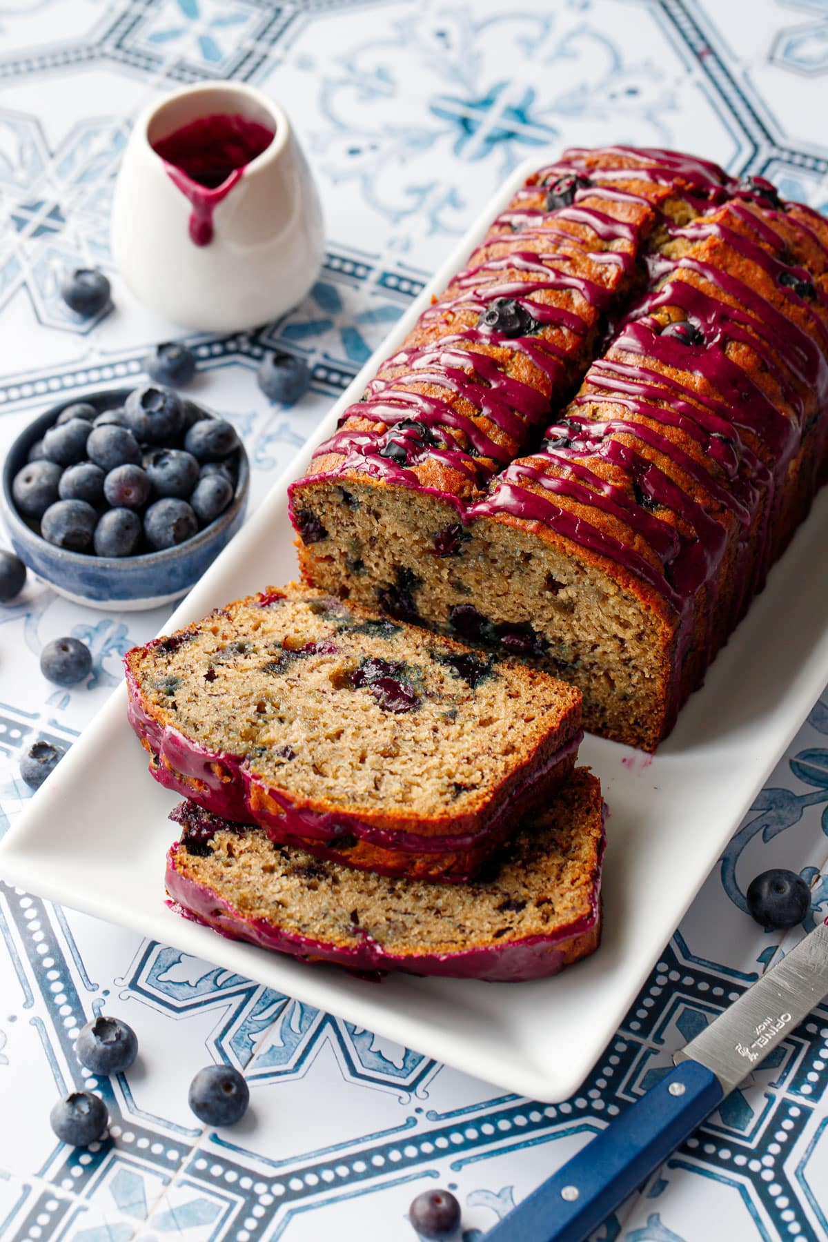 Loaf of Blueberry Banana Bread on a white rectangular plate with two slices lying down on a blue and white tile background.