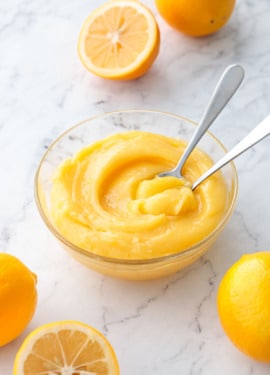 Glass bowl filled with a swirl of Meyer Lemon Curd and two spoons, with fresh lemons scattered around.