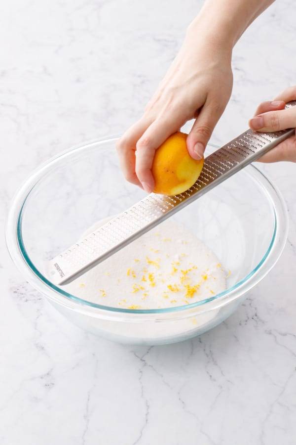 Zesting a Meyer lemon with a microplane grater over a bowl of granulated sugar.