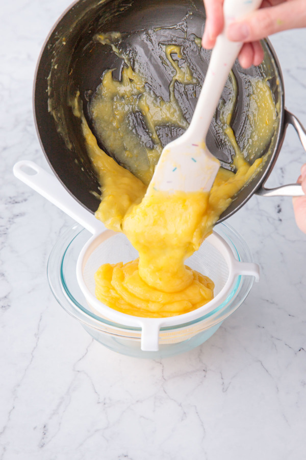 Pouring cooked lemon curd through a fine mesh sieve into a glass mixing bowl.