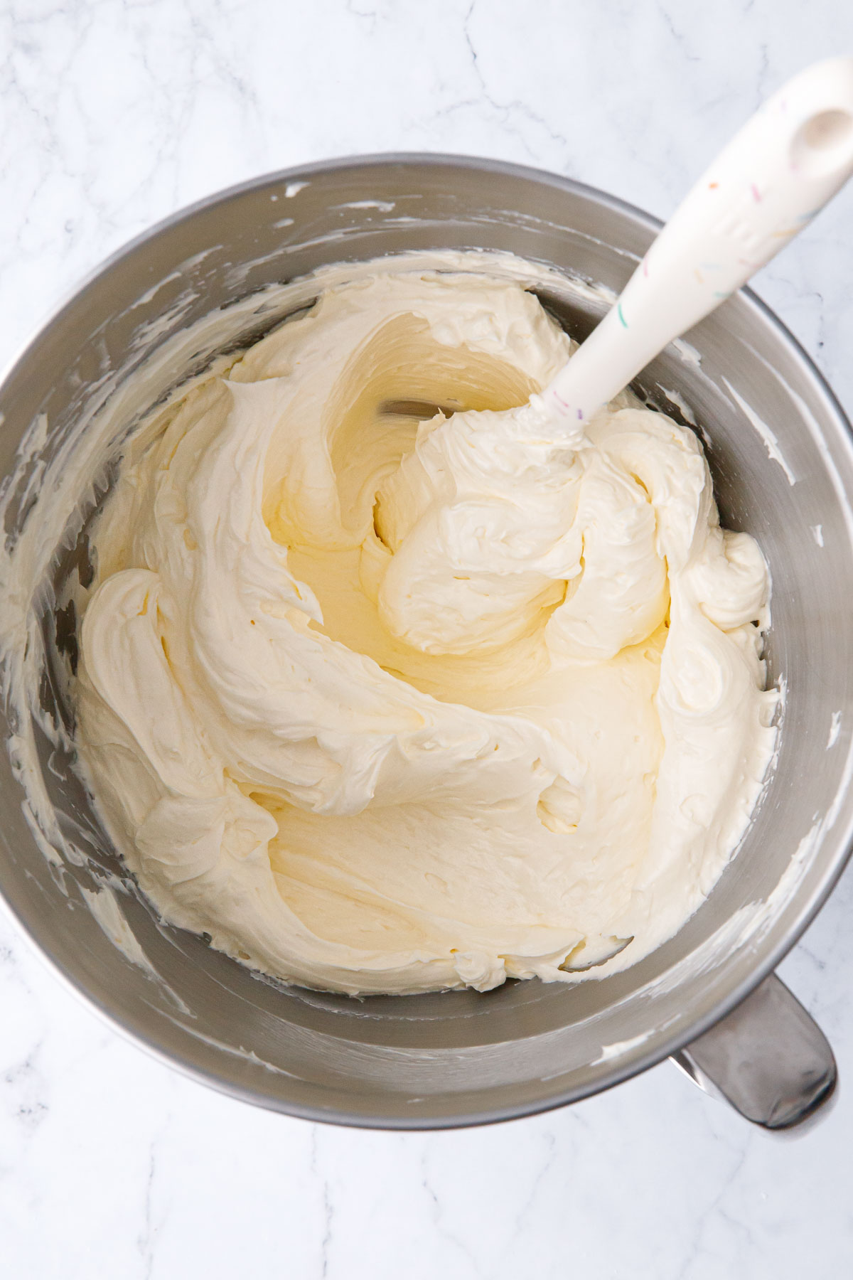 Bowl of Swiss Meringue Buttercream in a metal mixing bowl with white sprinkle spatula.