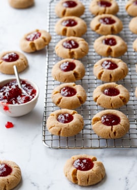 Rows of Peanut Butter & Jam Thumbprints on a wire baking rack, with bowl of strawberry jam and a few more cookies scattered around it.