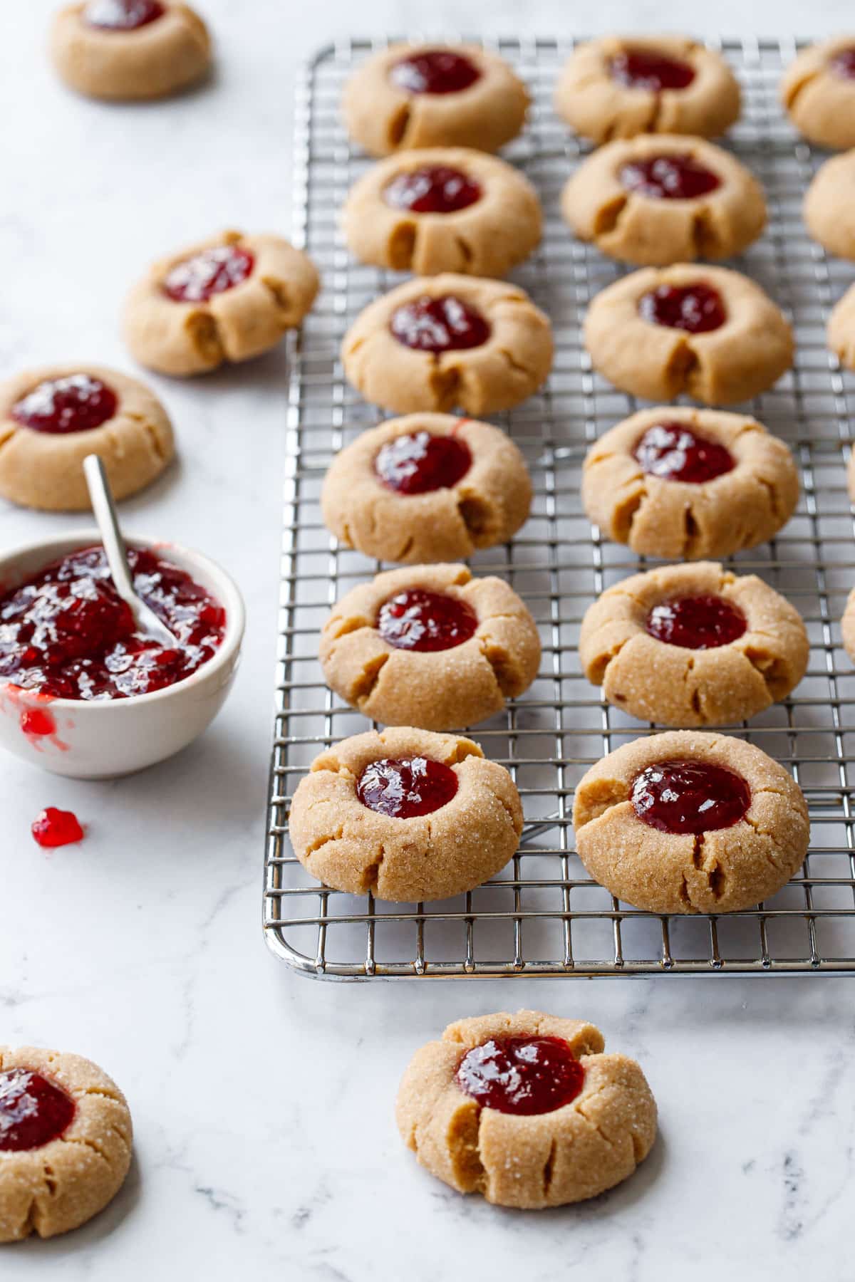 Rows of Peanut Butter & Jam Thumbprints on a wire baking rack, with bowl of strawberry jam and a few more cookies scattered around it.
