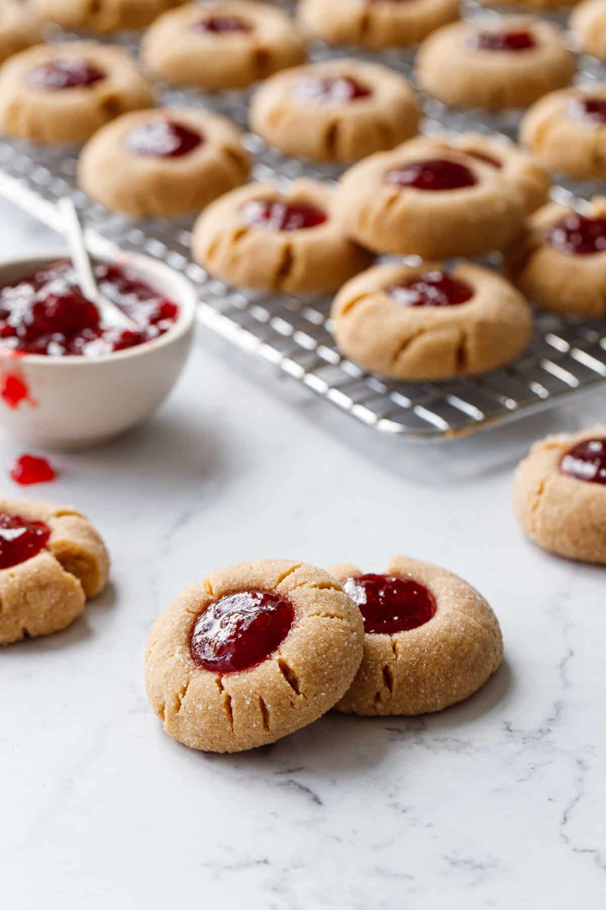 Peanut Butter & Jam Thumbprints on a marble background, with wire rack with more cookies plus a bowl of strawberry jam in the background.