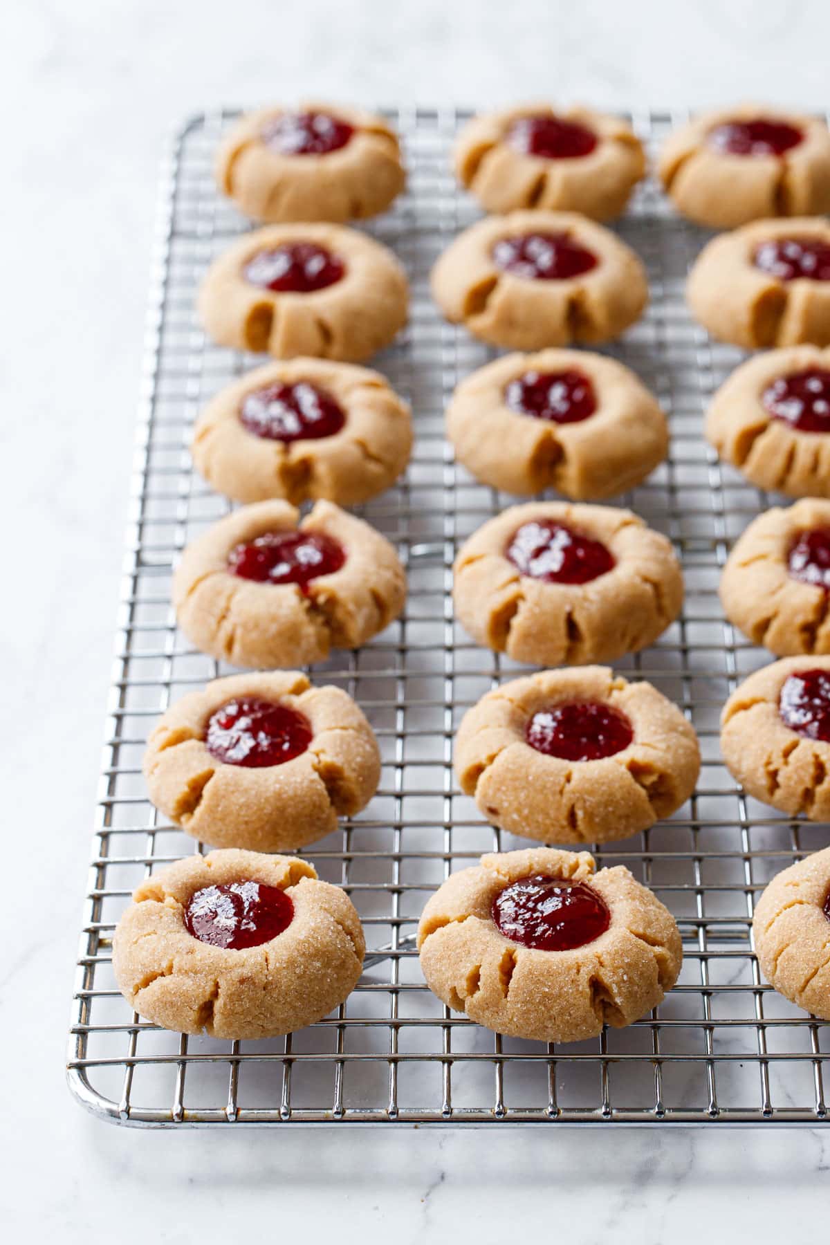 Rows of Peanut Butter & Jam Thumbprints on a wire baking rack, backlit to make the jam filling shine.
