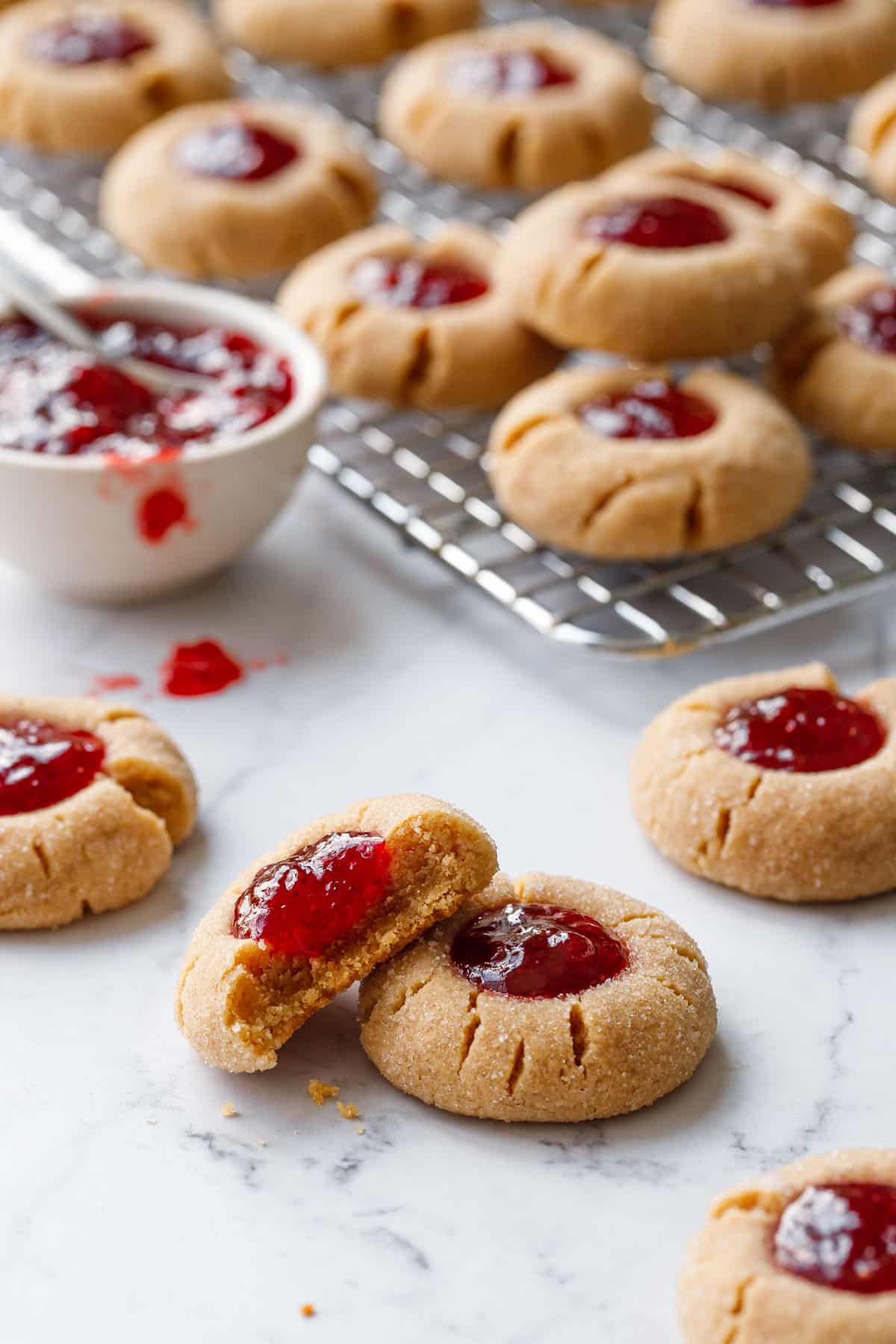 Peanut Butter & Jam Thumbprints on a marble background, one cookie with a bite taken out of it and more cookies plus a bowl of strawberry jam in the background.