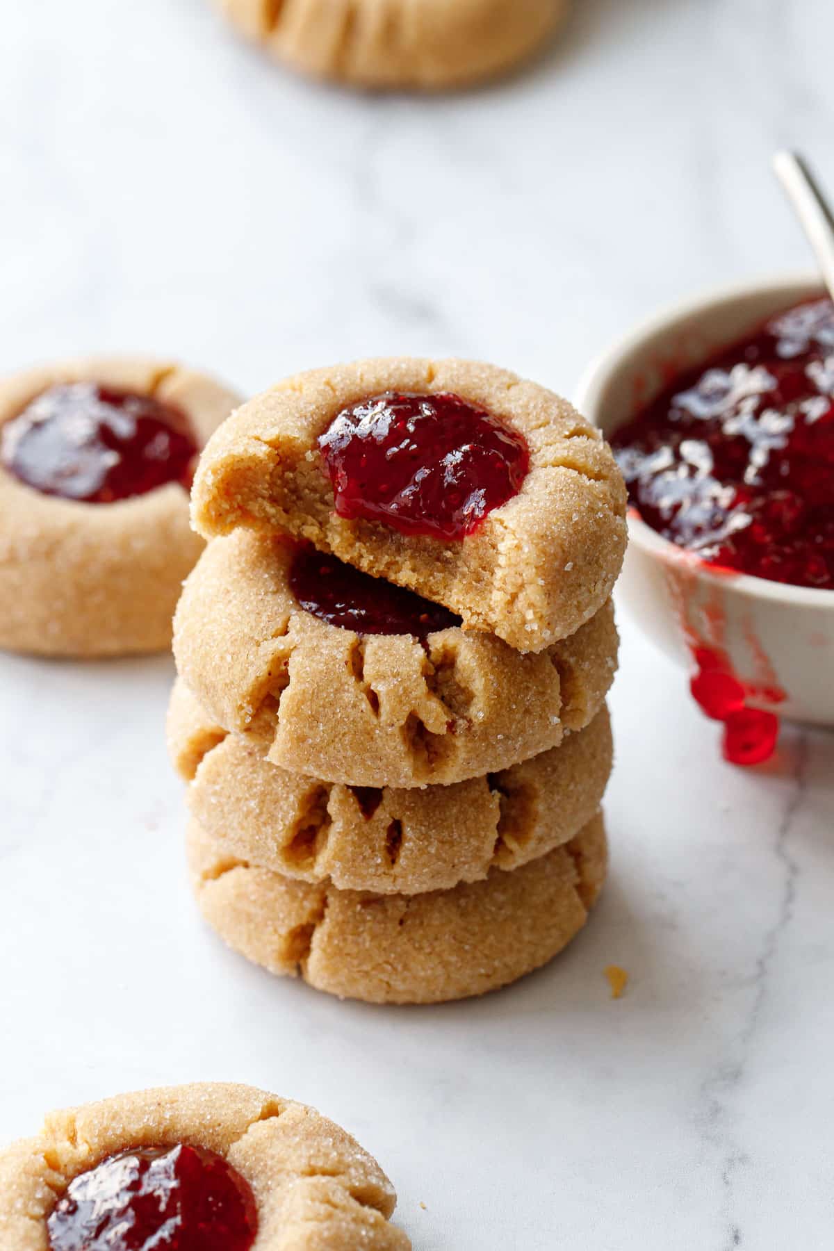 Stack of Peanut Butter & Jam Thumbprints, top cookie with a bite out of it, plus a bowl of strawberry jam on a marble background.
