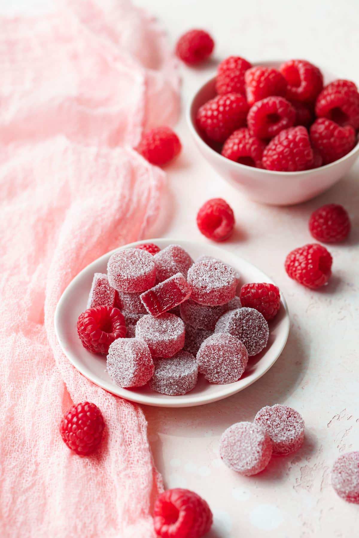 Small dish of Homemade Raspberry Pectin Gummies on a light pink background with linen and bowl of raspberries in the background.