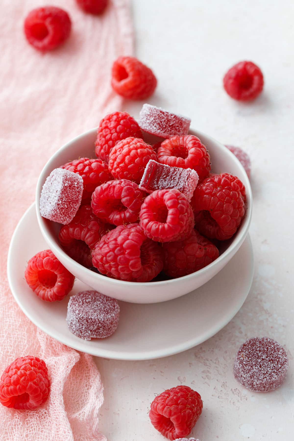 Bowl of fresh raspberries with a few Raspberry Gummy candies sitting on top and scattered around, with a light pink background and linen.