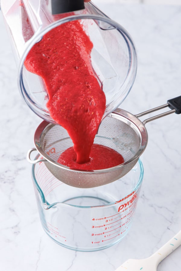 Pouring freshly blended raspberry puree from a blender through a fine mesh sieve into a spouted measuring cup.