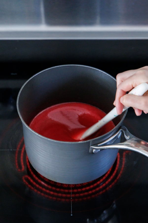 Stirring raspberry puree with sugar and corn syrup in a saucepan on the stovetop.