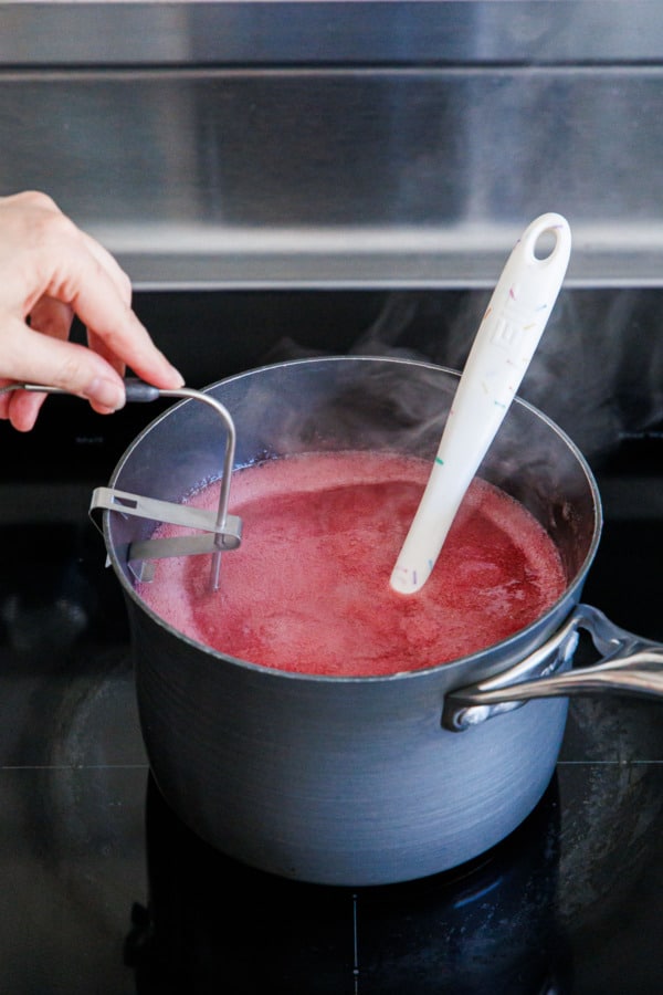 Affixing an instant read thermometer to the side of the saucepan filled with the simmering raspberry candy mixture.