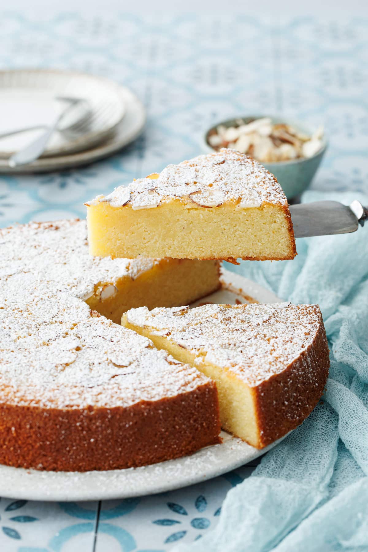 Slice of Flourless Almond Flour Cake lifted by a cake knife, on a blue tile background with linen napkin.