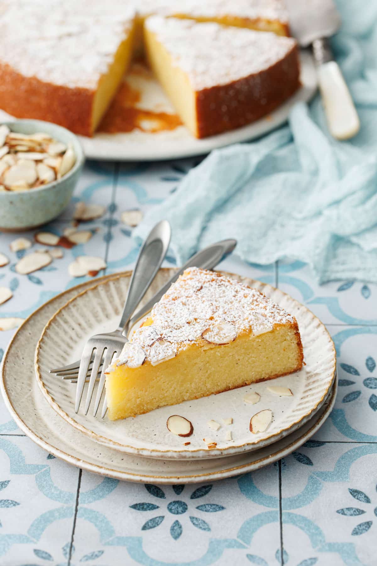Slice of Flourless Almond Flour Cake on a dessert plate with forks, rest of cake and bowl of sliced almonds in the background.