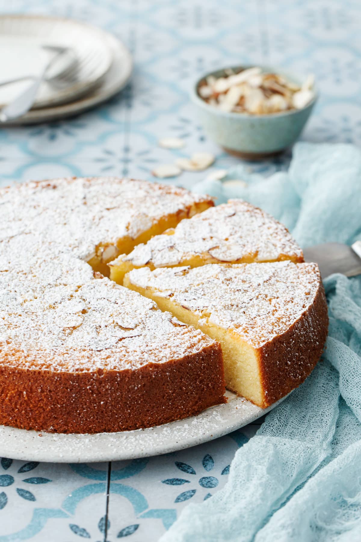 Flourless Almond Flour Cake, topped with sliced almonds and dusted with sugar, on a flat cake plate with two cut slices.