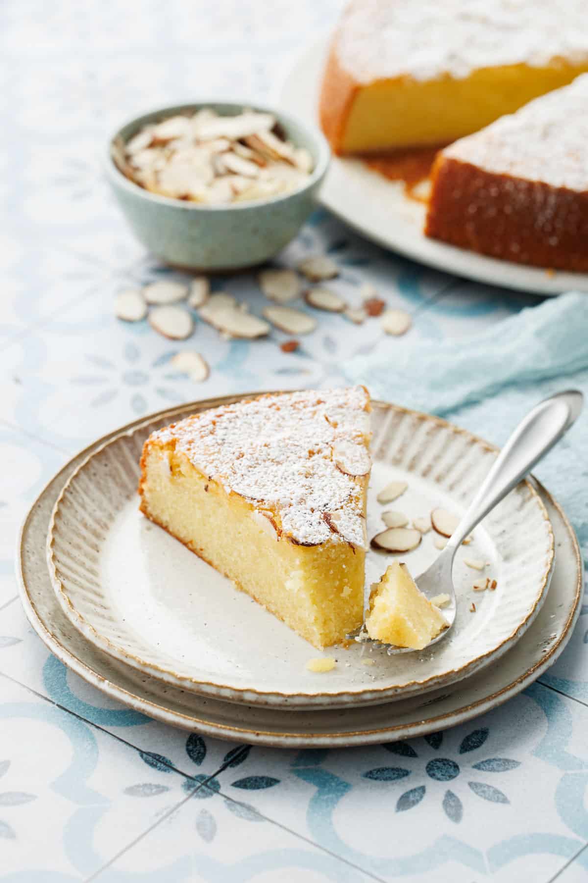 Slice of Flourless Almond Flour Cake on a dessert plate with a bite on a fork, rest of cake and bowl of sliced almonds in the background.