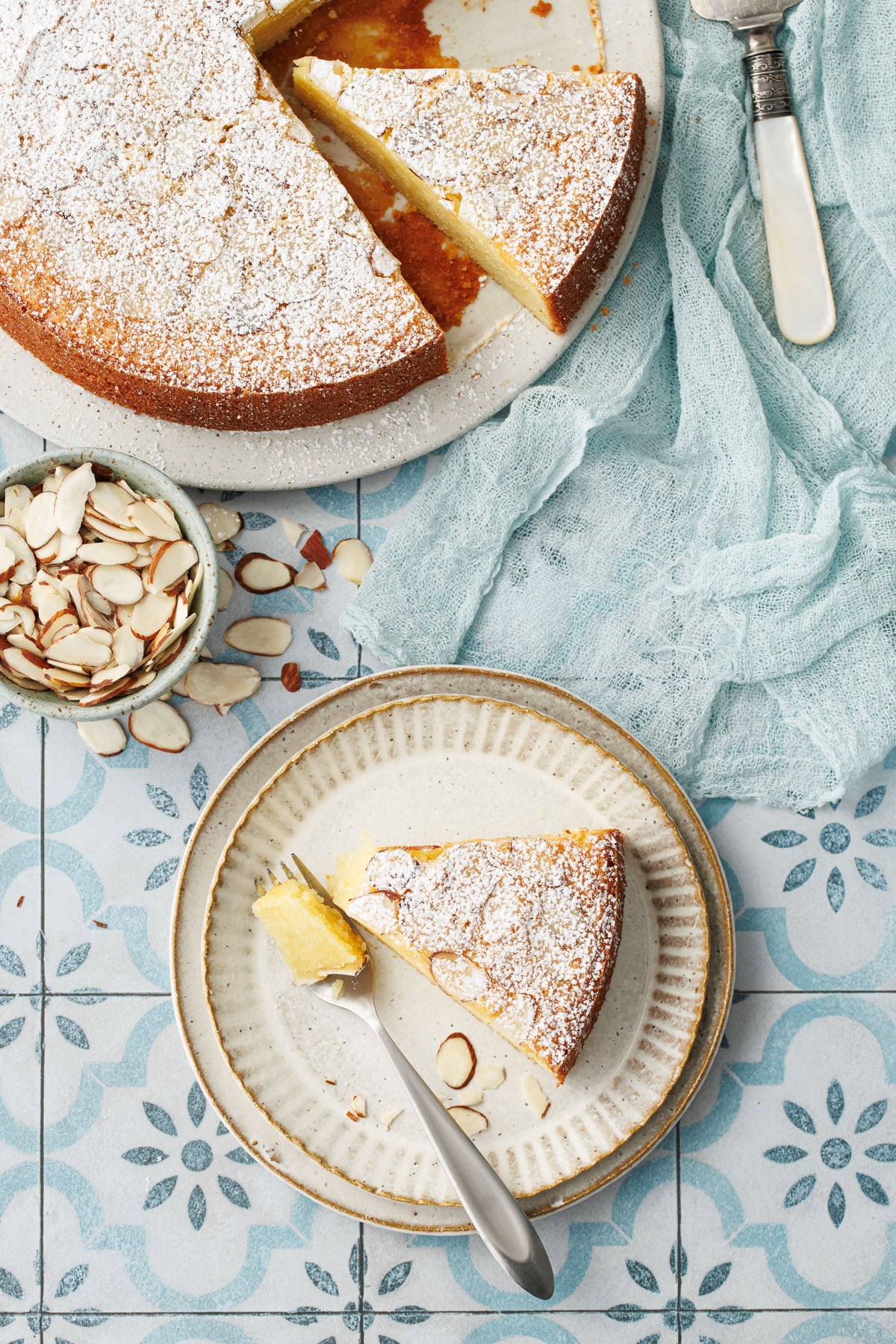 Overhead, slice Flourless Almond Flour Cake on a dessert plate with a fork, plus bowl of sliced almonds and partially sliced cake all on a blue tile background with light blue linen napkin.