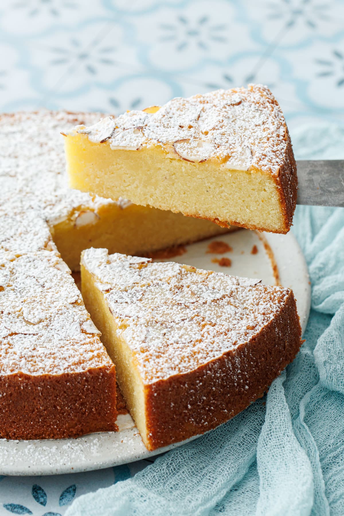 Slice of Flourless Almond Flour Cake lifted by a cake knife, on a blue tile background with linen napkin.