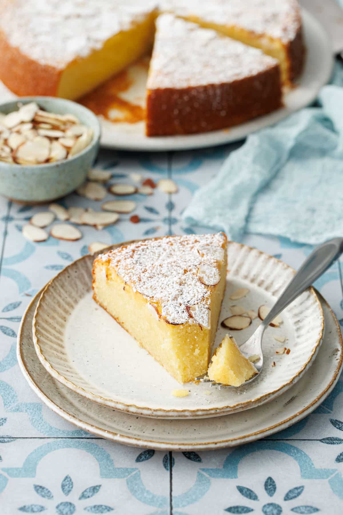 Slice of Flourless Almond Flour Cake on a dessert plate with a bite on a fork, rest of cake and bowl of sliced almonds in the background.