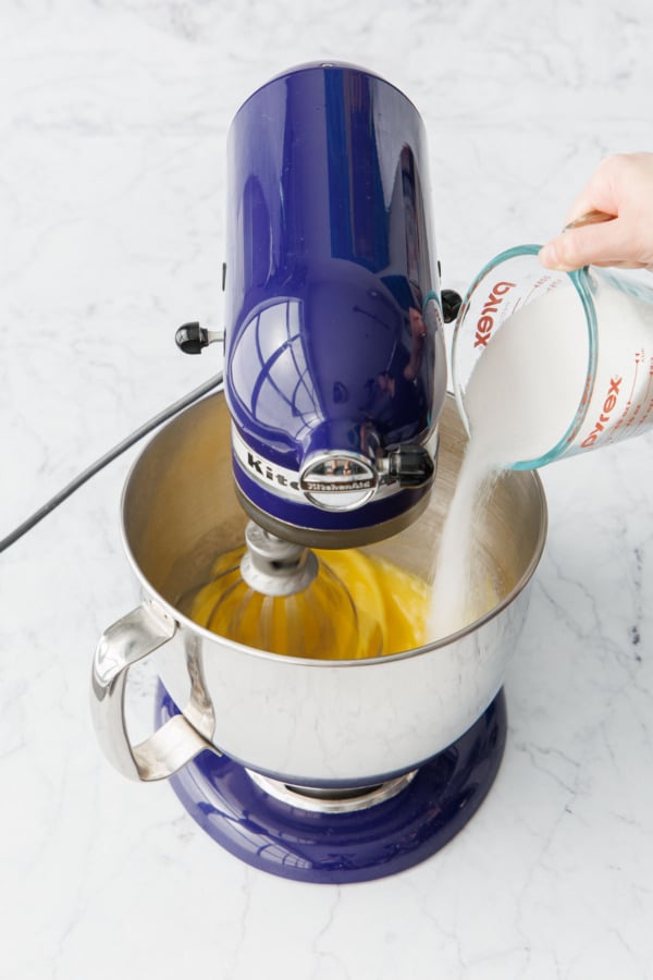 Pouring sugar into whole eggs in a stand mixer bowl fitted with the whisk attachment.