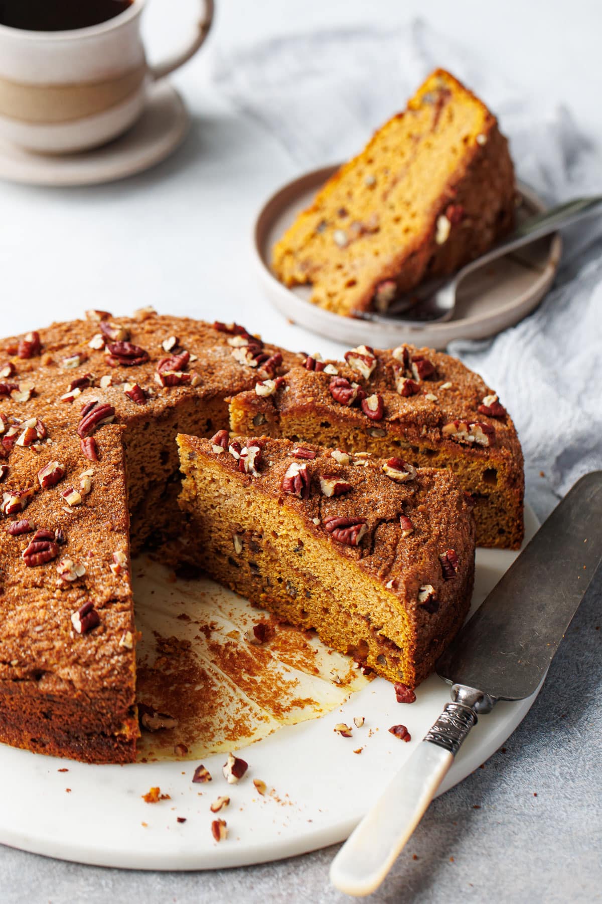 Triangular slices of Pumpkin Pecan Coffee Cake on a marble cake plate, one slice on a plate in the background with a cup of coffee.
