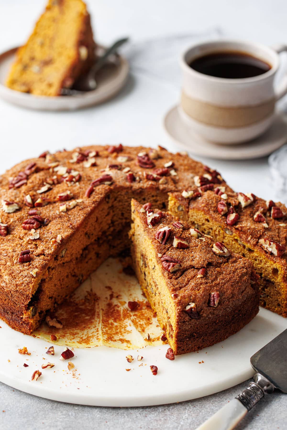Triangular slices of Pumpkin Pecan Coffee Cake on a marble cake plate, one slice on a plate in the background with a cup of coffee.