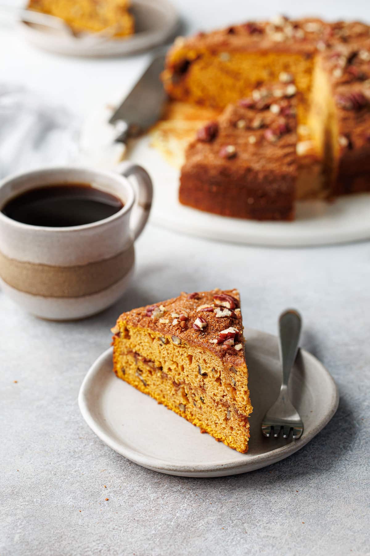 Triangular slice of Pumpkin Pecan Coffee Cake on a ceramic plate, with mug of coffee and the rest of the sliced cake in the background.