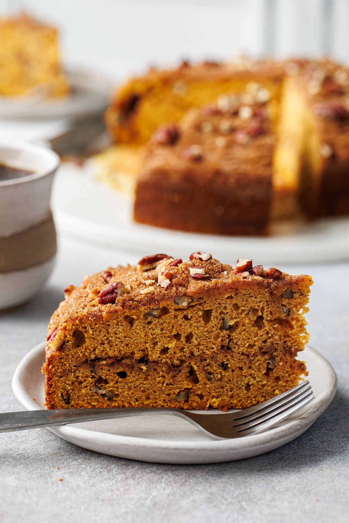 Slice of Pumpkin Pecan Coffee Cake on a ceramic plate, coffee mug and remaining cake in the background.